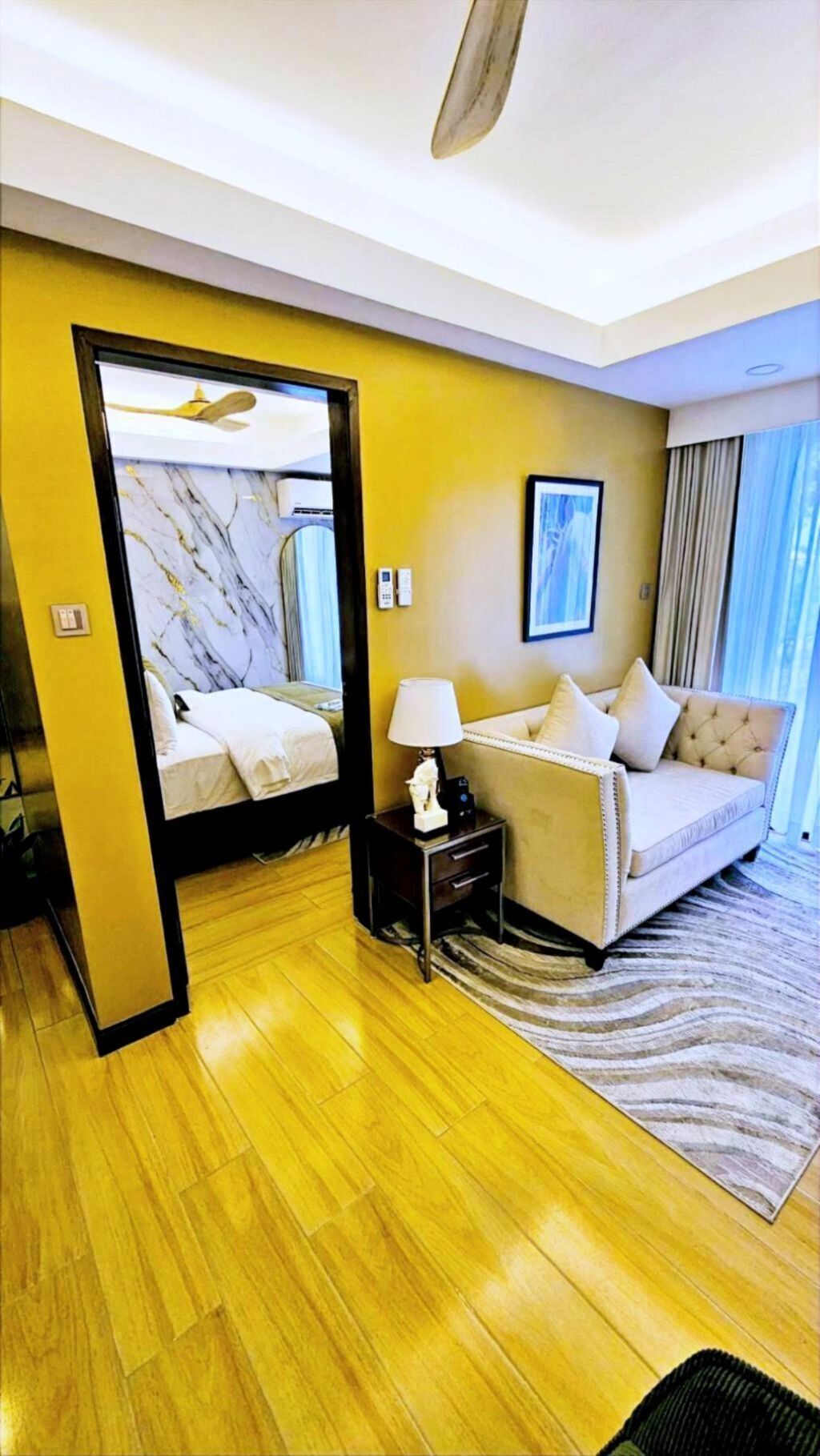A hotel room with a bed, a sofa, a nightstand with a lamp, a mirror, and a window with curtains.