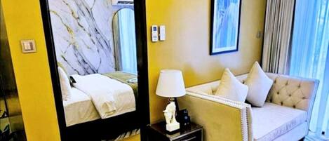 A hotel room with a bed, a sofa, a nightstand with a lamp, a mirror, and a window with curtains.