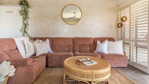 Een woonkamer met een roze zithoek, een rond rieten bijzettafeltje, een spiegel aan de muur en een hangplant aan het plafond.