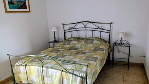 Ein Einzelbett mit Metallgestell, eine grün gemusterte Bettdecke, zwei Nachttische mit Lampen und ein gerahmtes Bild an der Wand.