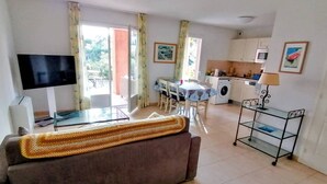 Ein Wohnzimmer mit Sofa, Fernseher, Essbereich mit Tisch und Stühlen, Küche mit Schränken und Waschmaschine sowie ein Balkon mit Vorhängen.