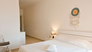 A neatly made bed with white linens, a bedside lamp, and a wall hanging. - Il-qastna Holiday House-zejtun (Zejtun)