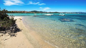 Une plage à l’eau turquoise et limpide, un rivage sablonneux et une vue lointaine sur une ville côtière avec ses bâtiments.