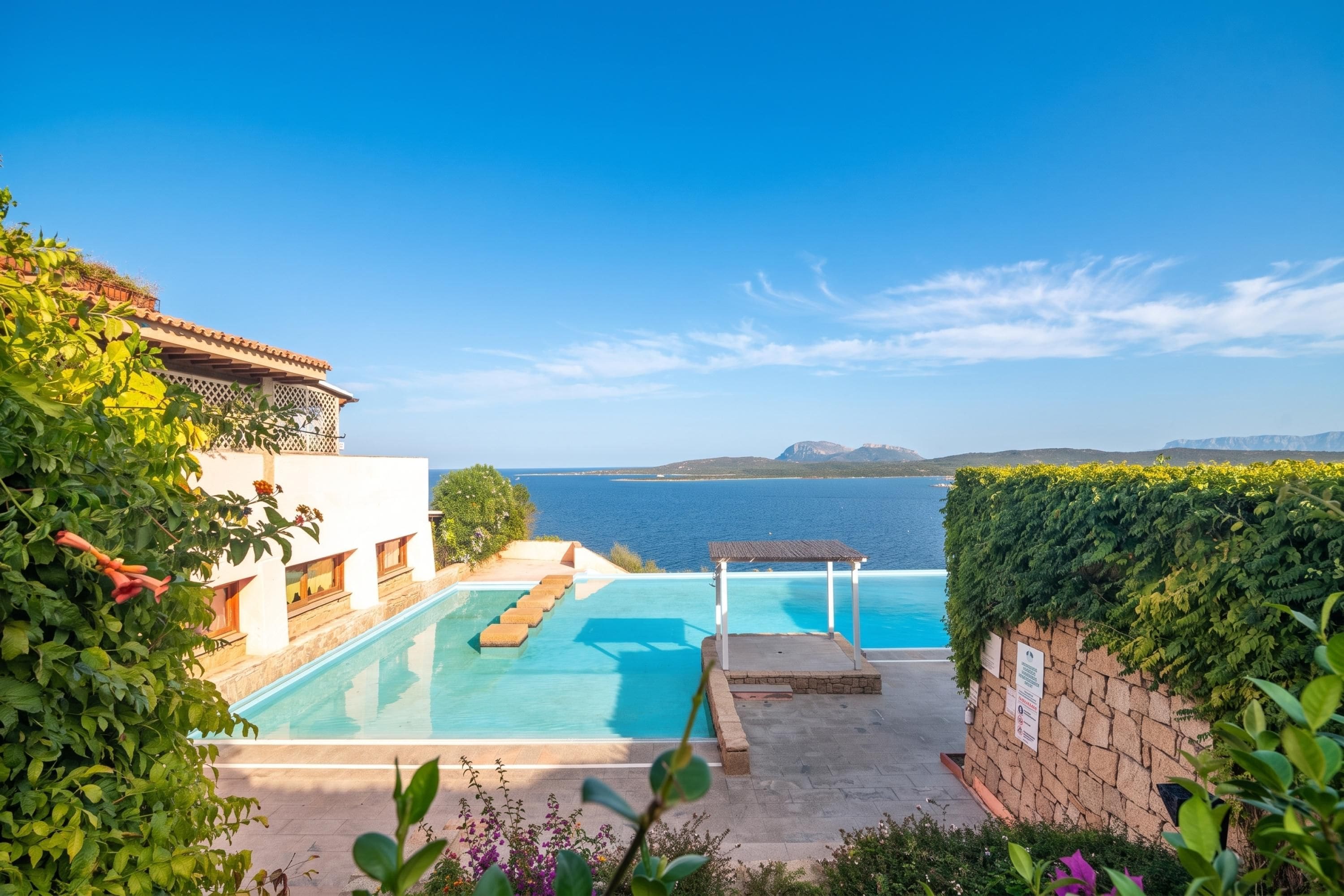 A pool area with a view of the sea, surrounded by greenery and a building with a tiled roof.