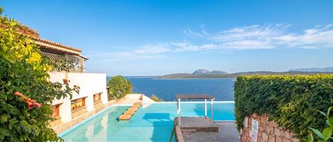 A pool area with a view of the sea, surrounded by greenery and a building with a tiled roof.