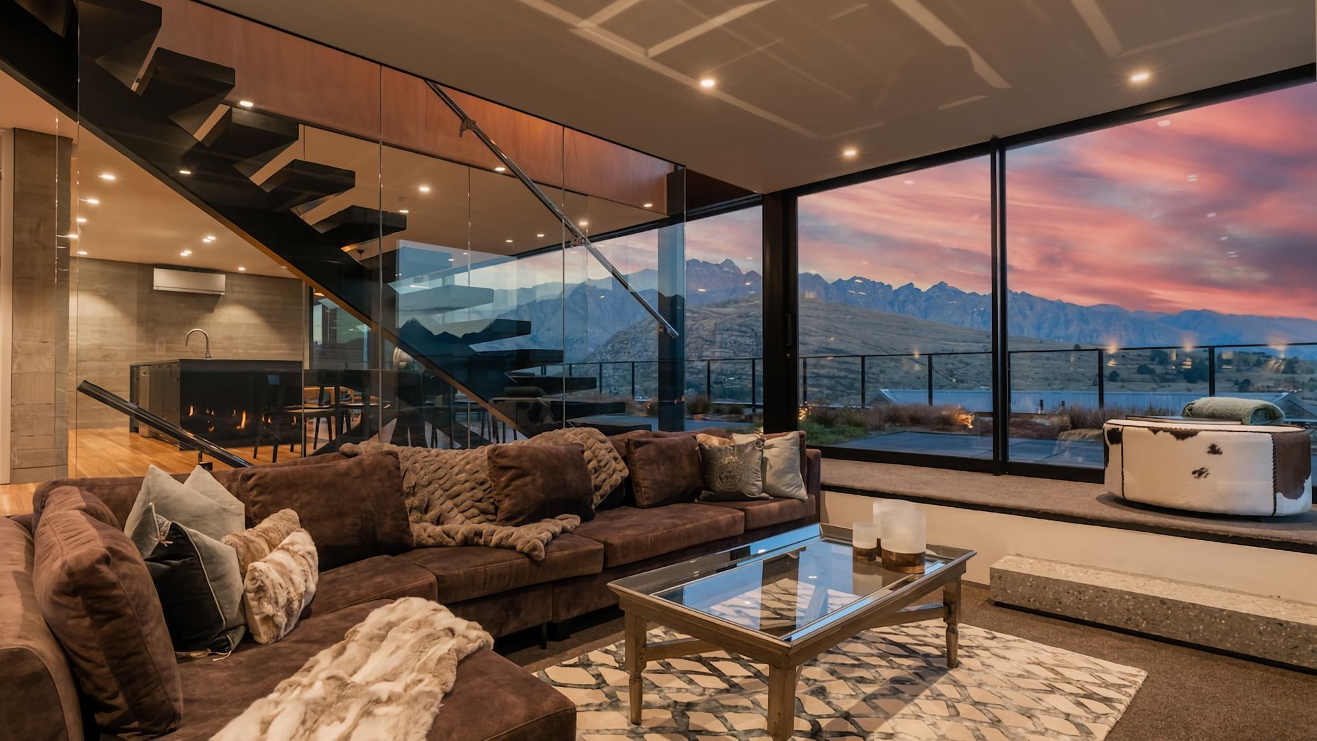 A modern living room with a large glass window offering a mountain view, a cozy brown sofa, a glass coffee table, and a staircase leading to an upper level.