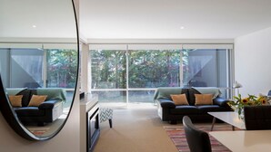 A modern living room with two black sofas, a coffee table, and a dining table with a vase of sunflowers. Large windows offer a view of greenery outside. - Apartment G3 3BR Near Queenstown Centre (Queenstown)