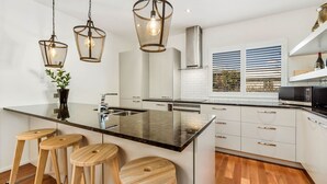 A modern kitchen with a central island, bar stools, pendant lighting, and a view of the outdoors through a window with blinds. - Central Queenstown 3 Bed Villa (Queenstown)