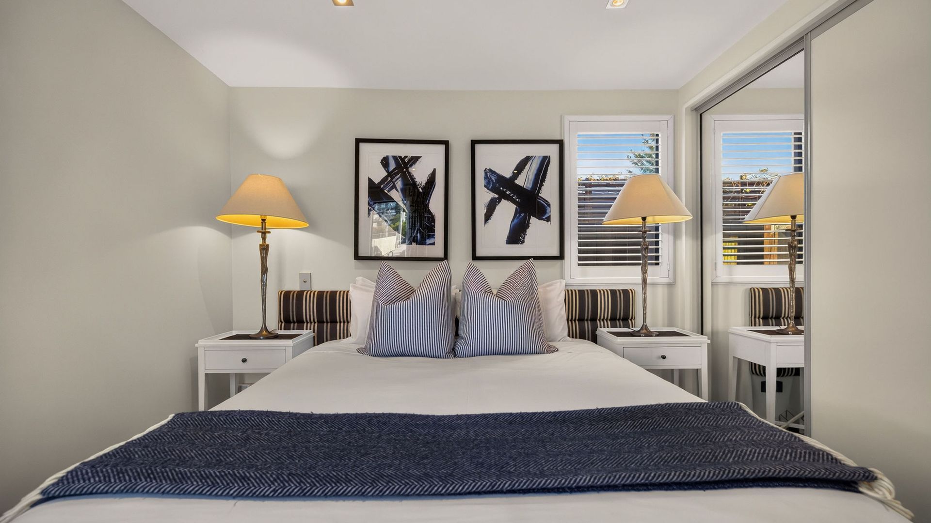 Een moderne hotelkamer met een groot bed, twee nachtkastjes, een raam met zonwering en twee ingelijste abstracte schilderijen.