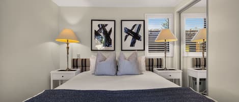 A modern hotel room with a large bed, two bedside tables, a window with blinds, and two framed abstract paintings.