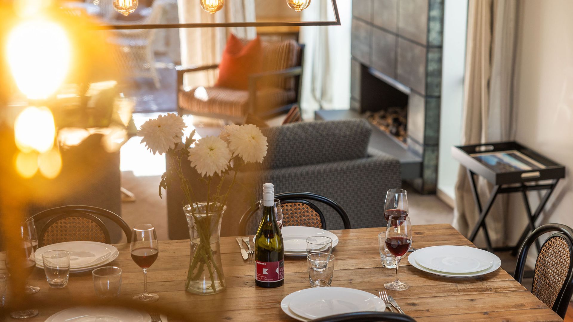 A dining table set with plates, wine glasses, and a bottle of wine, with a vase of flowers and a fireplace in the background.