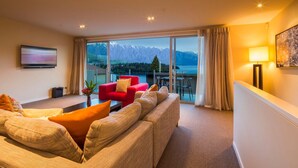 Een moderne hotelkamer met een groot raam met uitzicht op bergen en een meer. Er staat een rode fauteuil, een beige bank en een flatscreen televisie aan de muur.