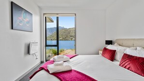 Quarto de hotel com uma cama grande, vista para um lago e montanhas, janela com vista e um quadro na parede.