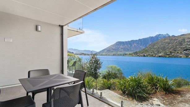 Varanda com mesa e cadeiras, vista para um lago com montanhas ao fundo.