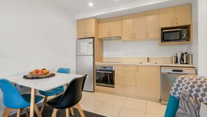 Une cuisine compacte avec des meubles de couleur bois clair, un four à micro-ondes, un réfrigérateur et un lave-vaisselle. On y trouve une table à manger avec des chaises et un panier de fruits posé dessus.