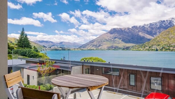 Un balcon avec une table et des chaises en bois, donnant sur un lac avec des montagnes en arrière-plan.