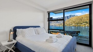 Une chambre à coucher avec un lit, des oreillers, une table de chevet, une lampe et une vue sur un lac et des montagnes, visible à travers de grandes fenêtres.