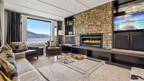 A modern living room with a stone fireplace, comfortable seating, a large window with a view of mountains, and a flat-screen TV mounted on the wall. - Commonage Grand Villa 2 Bedroom (Queenstown)