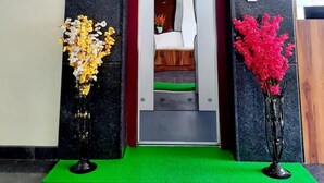A hotel reception area with a modern design, featuring a reception desk, a staircase leading to the upper floor, and two decorative flower arrangements on green mats. - Hotel Royal Suites (Bengaluru)