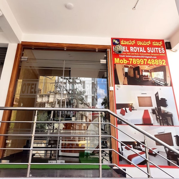 A hotel entrance with a glass facade, a staircase leading to the entrance, and a signboard advertising 'EL ROYAL SUITES' with contact information. - Hotel Royal Suites (Bengaluru)