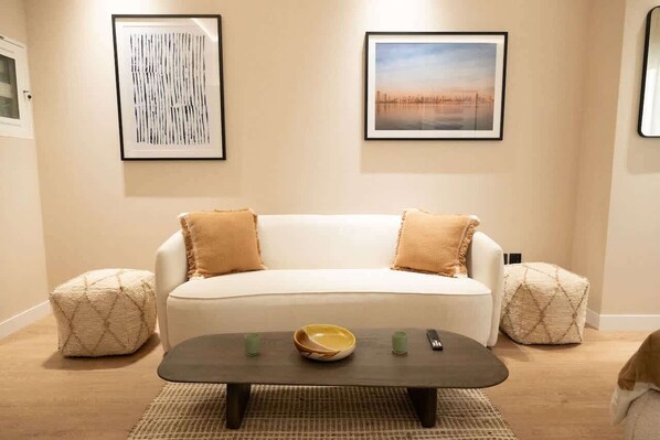 A living room with a white sofa, two beige ottomans, a dark coffee table with a bowl and remote controls, and two framed pictures on the wall. - Chic studio located in the heart of Business Bay (buisness bay)