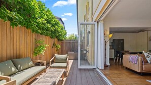 Une terrasse dotée de meubles en bois, une porte-fenêtre coulissante donnant sur un espace intérieur avec une table et des chaises.
