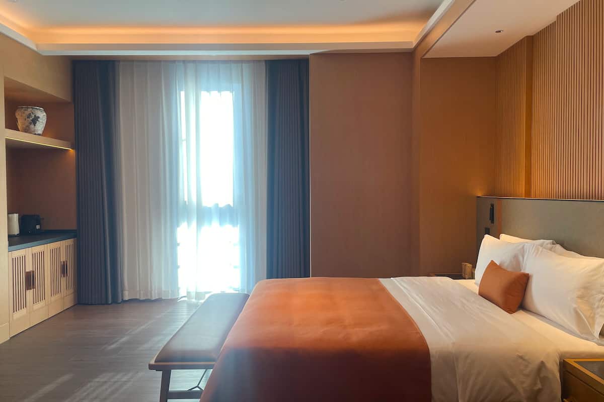 Habitación de hotel con una cama grande, una mesita de noche, un banco, un armario pequeño y una ventana con cortinas.