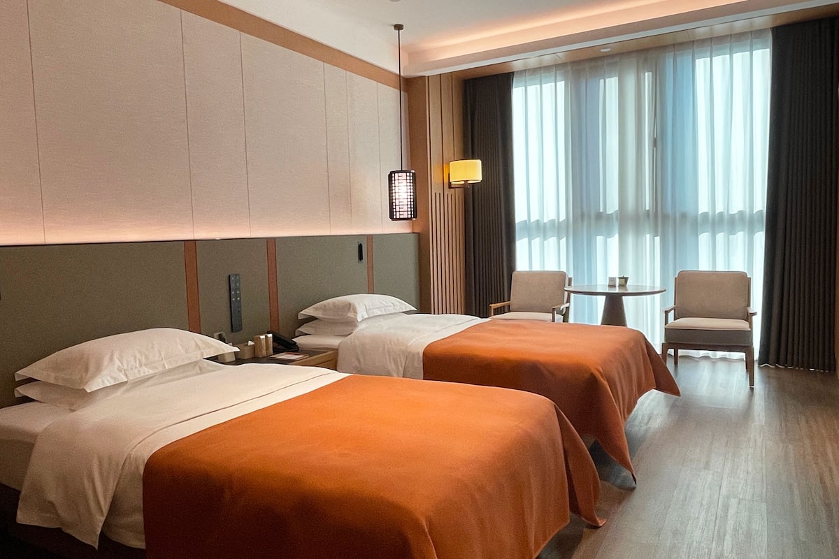 Habitación de hotel con dos camas, una mesita pequeña, dos sillas y un ventanal grande con cortinas.