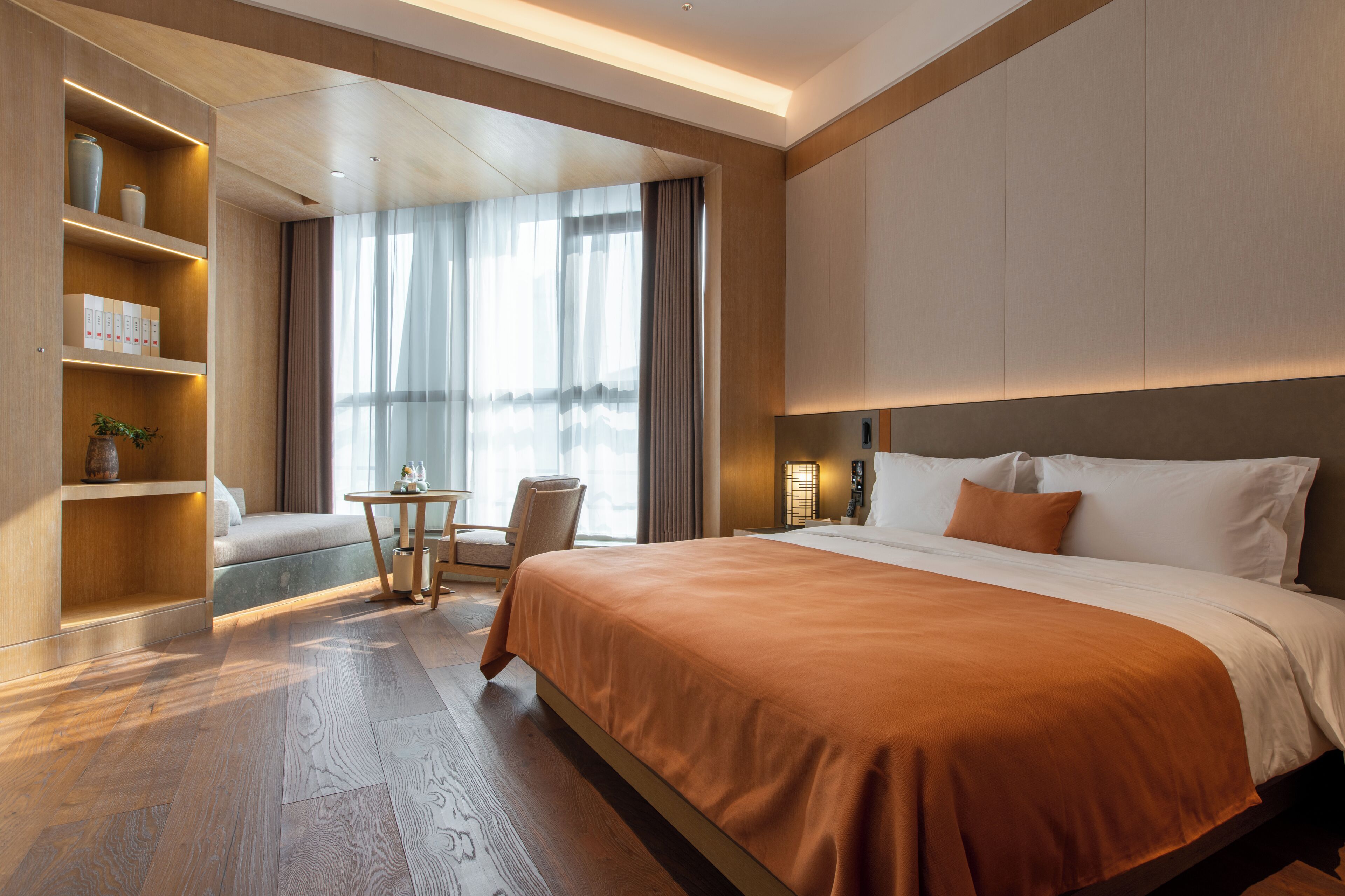 Een moderne hotelkamer met een groot bed, een zithoek met een tafel en stoelen, en ingebouwde planken met decoratieve objecten.