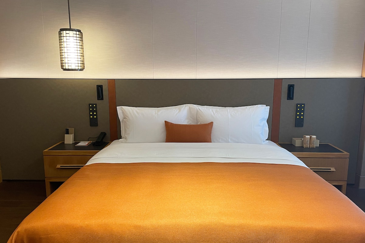 Habitación de hotel con una cama grande, una mesita de noche con un teléfono y una lámpara, y un panel de control en la pared.