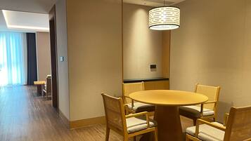 Habitación de hotel con una mesa redonda y cuatro sillas, una lámpara colgante y vistas a un pasillo con una cama y una ventana.