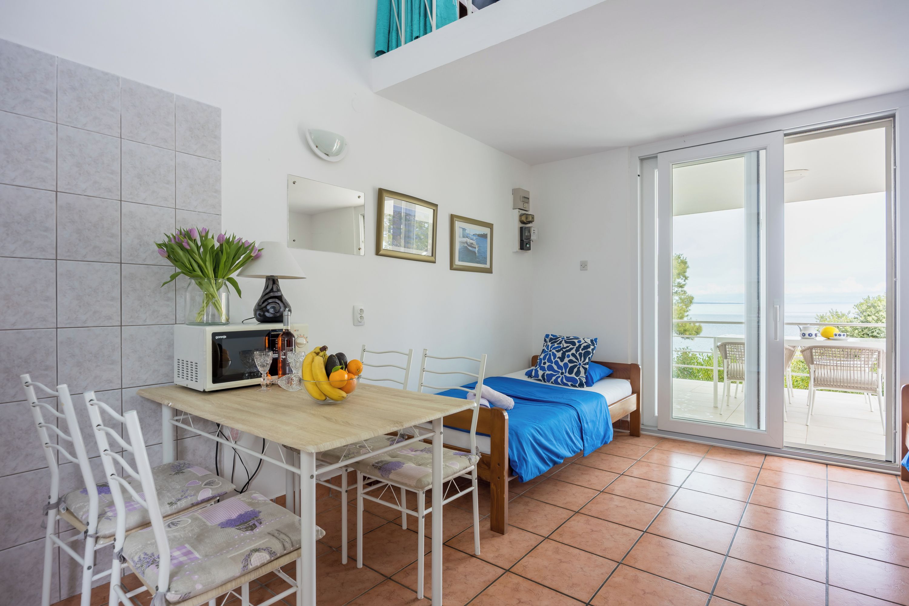 Een kamer met een eettafel, stoelen, een bed met een blauwe dekbedovertrek, een balkon met uitzicht en een magnetron op de tafel.