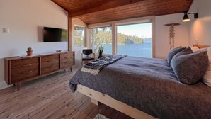 A bedroom with a wooden ceiling, a bed with grey bedding, a wooden dresser, a TV, and a view of a lake. - Marielle’s Lookout -  Your Oceanfront Escape (Tofino)
