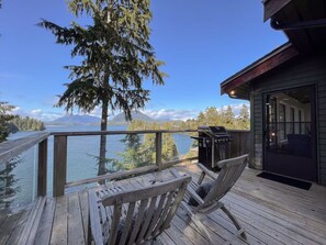 A wooden deck with a view of a lake and mountains, a grill, and a glass door leading to an interior space.