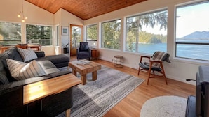 A spacious living room with a wooden ceiling, large windows offering a view of a lake and mountains, a sofa, a coffee table, and a chair. - Marielle’s Lookout -  Your Oceanfront Escape (Tofino)