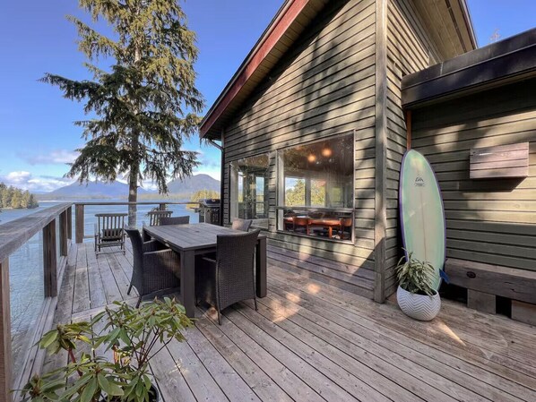 A wooden deck with outdoor furniture, a surfboard, and a view of a lake and mountains.