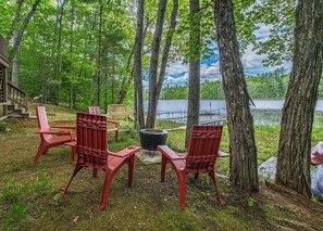 En træterrasse ved søen med røde Adirondack-stole, en bålplads og en træbænk.