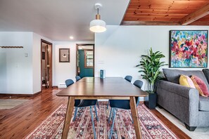 Un coin repas avec une table en bois, des chaises bleues, un tapis à motifs, un canapé gris avec des coussins, une plante en pot et un tableau abstrait coloré accroché au mur.