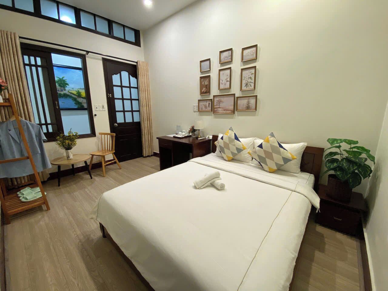 A hotel room with a large bed, a desk, a chair, a small table with a vase, and a window with curtains.