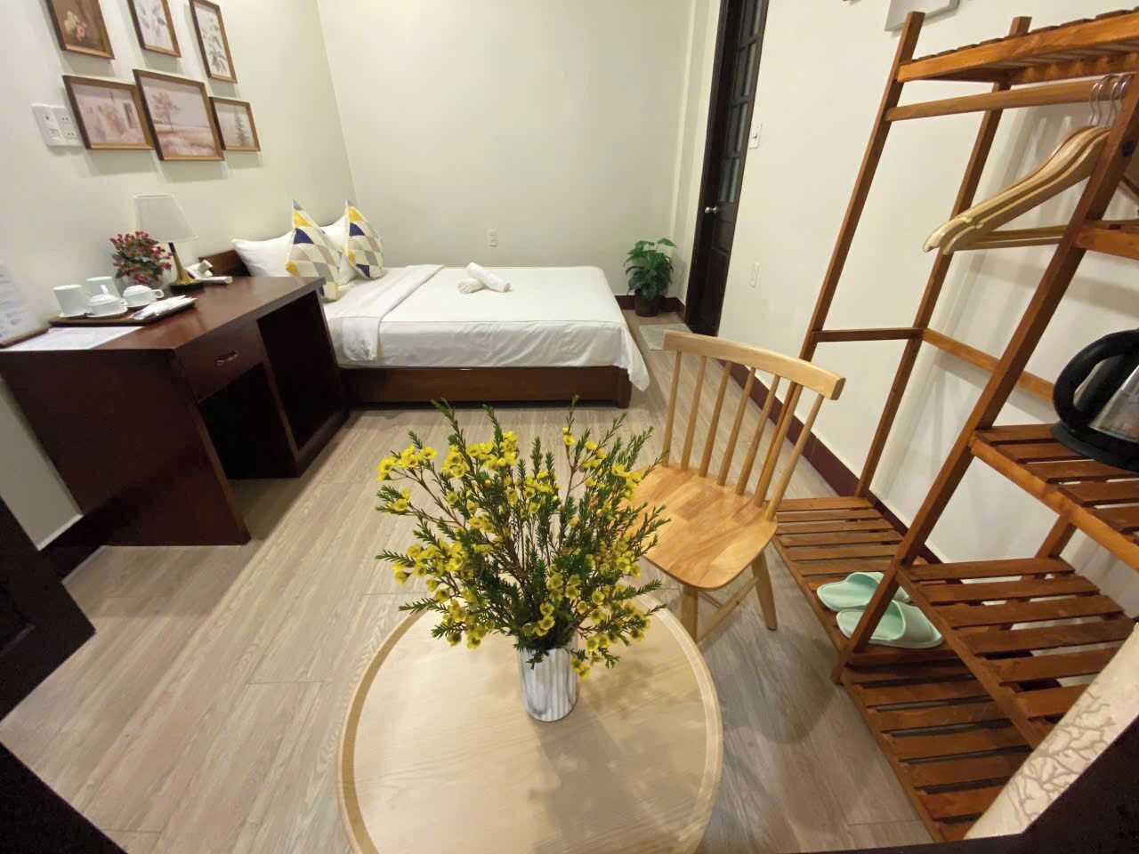 Une petite chambre d’hôtel avec un lit, un bureau, une chaise, une étagère en bois et un vase contenant des fleurs jaunes.