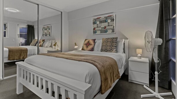 Ein modernes Schlafzimmer mit einem großen Bett, Nachttischen, einem Spiegel und einem Standventilator.