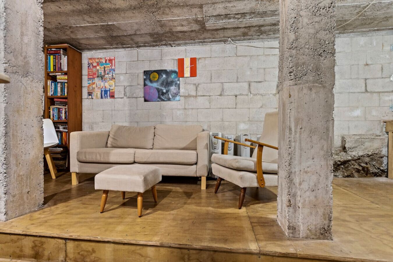 Un salon avec un canapé beige, un fauteuil en bois et une bibliothèque adossée à un mur en béton.