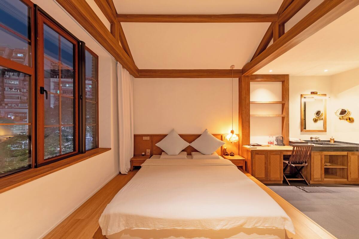 A hotel room with a large bed, wooden beams, a desk with a chair, a mirror, and a window with curtains.