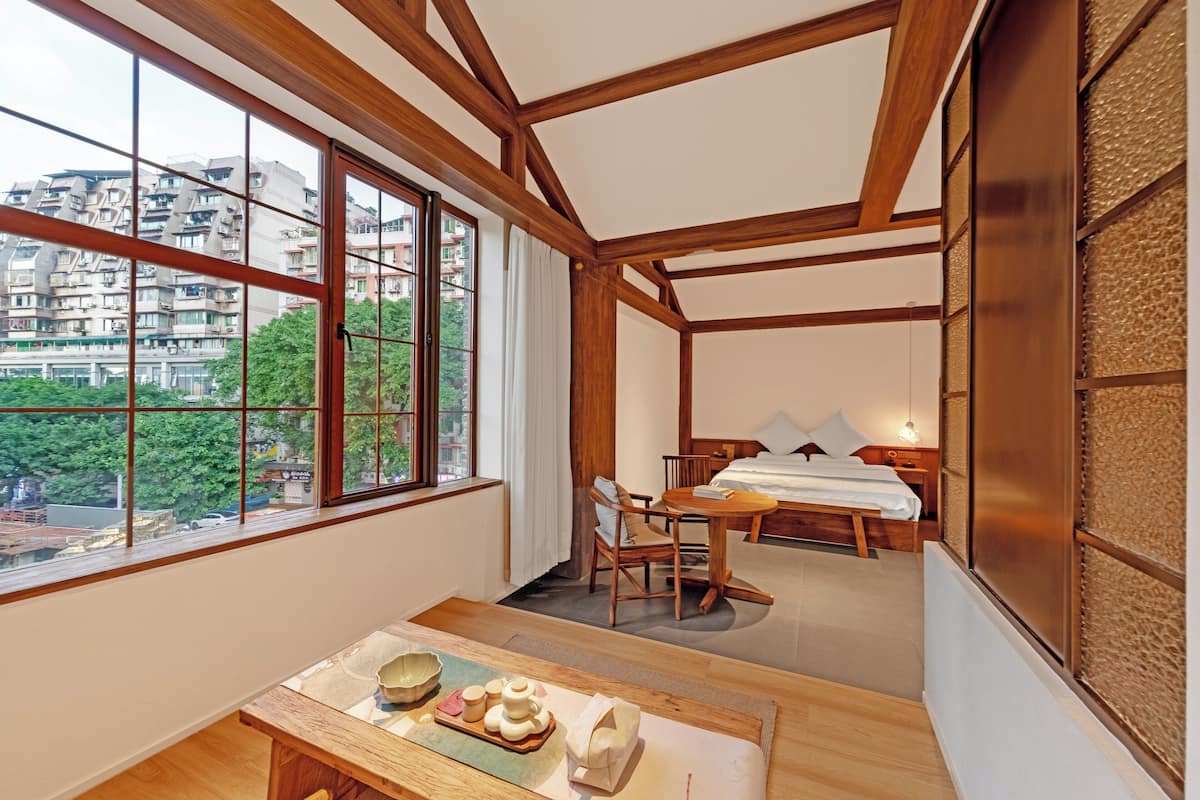 A hotel room with a large window, wooden beams, a dining table with a tray of items, and a bed with a wooden headboard.