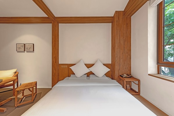 Un dormitorio moderno con cabecera de madera, una cama con sábanas blancas, una mesita de noche de madera y un ventanal con vistas a árboles.