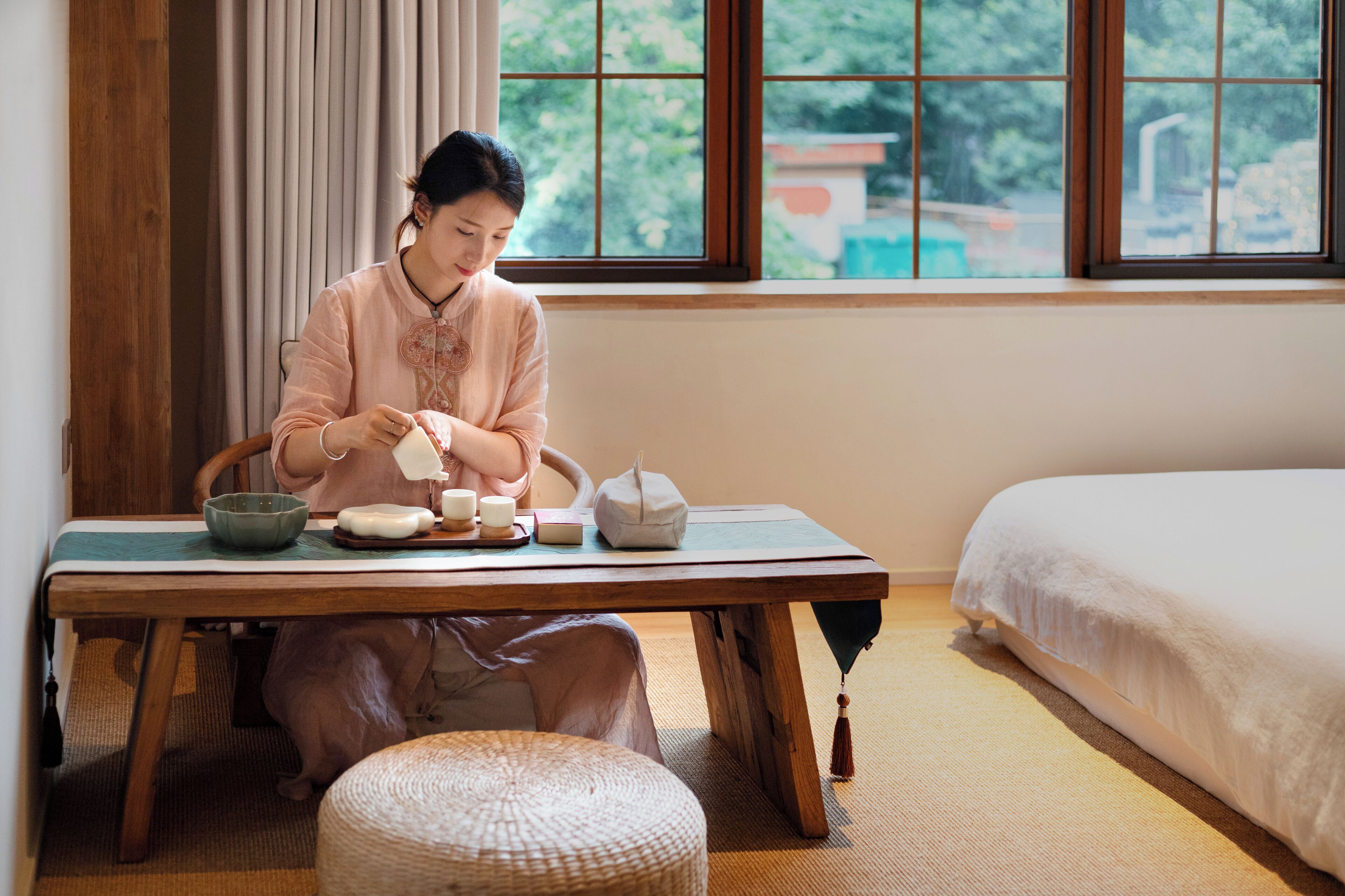 A room with a wooden table, a bed, and a window with a view of greenery outside.