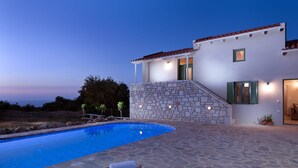 Area piscina con pavimento in pietra, una sedia a sdraio e un edificio con facciata in pietra e persiane verdi.