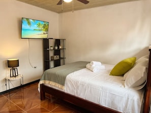 A hotel room with a bed, a nightstand, a lamp, a wall-mounted TV, and a shelf with decorative items.