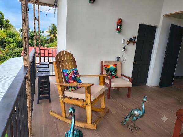 A wooden rocking chair with a colorful cushion, a matching chair, and two decorative peacock statues on a balcony with a view of greenery.