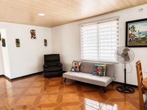 A room with a sofa, a chair, a standing fan, and a window with blinds.